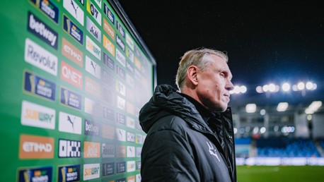 A person stands beside a backdrop covered in various sponsor logos with Etihad Stadium lights in the background.
