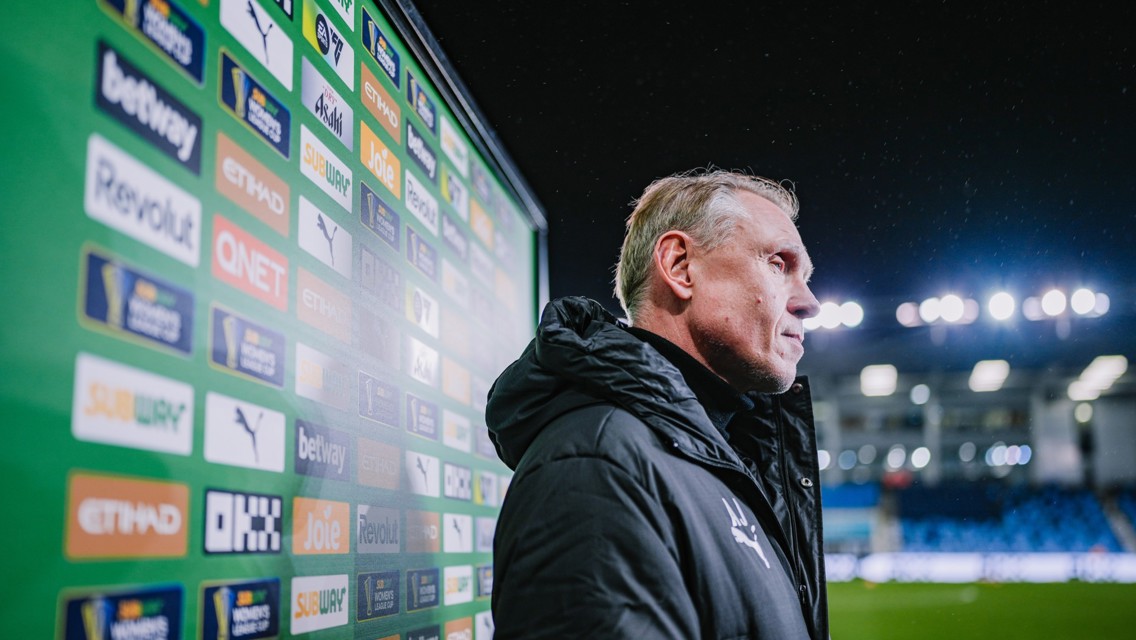A person stands beside a backdrop covered in various sponsor logos with Etihad Stadium lights in the background.