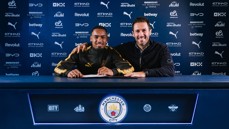 Two individuals smiling behind a table with Manchester City branding, likely involving a football contract signing.