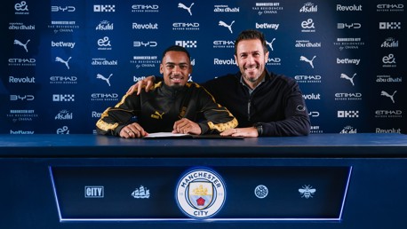 Two individuals smiling behind a table with Manchester City branding, likely involving a football contract signing.