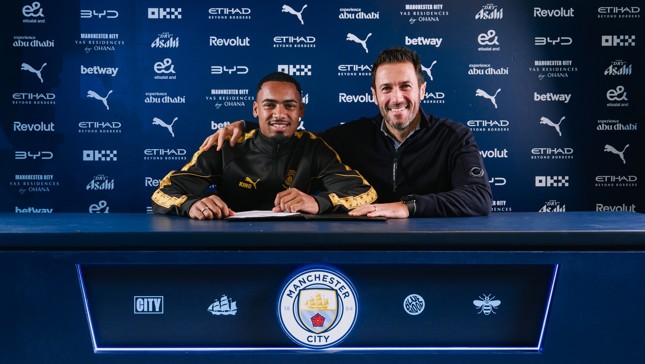 Two individuals smiling behind a table with Manchester City branding, likely involving a football contract signing.