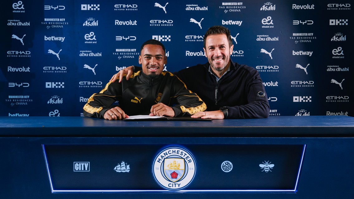 Two individuals smiling behind a table with Manchester City branding, likely involving a football contract signing.