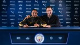 Two individuals smiling behind a table with Manchester City branding, likely involving a football contract signing.
