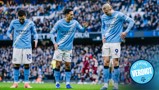 Three Manchester City football players in a team discussion on the field, with blurred faces and crowd in background.