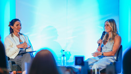 Two women seated on stage under blue lighting having a discussion. One is holding a microphone and the other holding a clipboard.