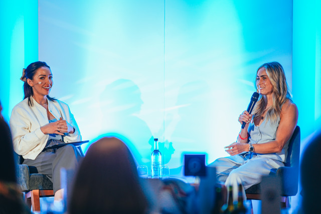 Two women seated on stage under blue lighting having a discussion. One is holding a microphone and the other holding a clipboard.