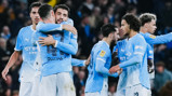 A group of Manchester City players in light blue kits, celebrating on a football field.