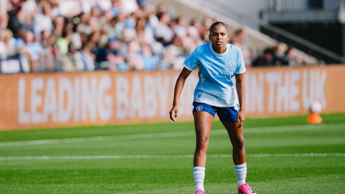A female soccer player wearing a light blue jersey and dark blue shorts on a soccer field during a game, with a blurred crowd in the background.