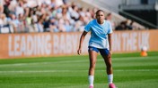 A female soccer player wearing a light blue jersey and dark blue shorts on a soccer field during a game, with a blurred crowd in the background.