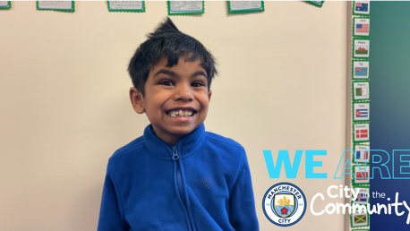 A young boy in a blue jacket is standing against a beige wall. There are small flags with country names on cards on the wall. The image features the Manchester City logo and text 'WE ARE City in the Community.'