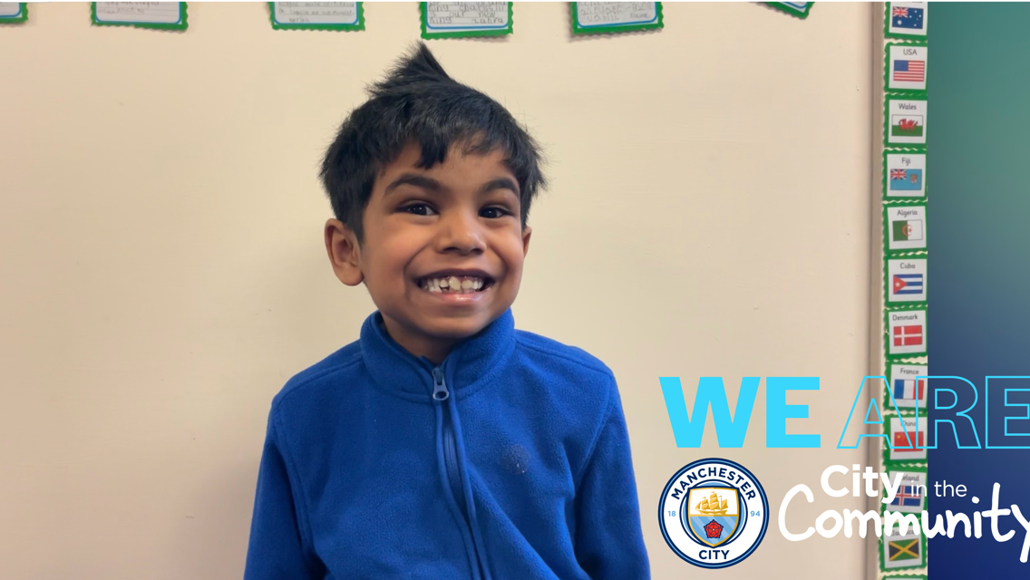 A young boy in a blue jacket is standing against a beige wall. There are small flags with country names on cards on the wall. The image features the Manchester City logo and text 'WE ARE City in the Community.'