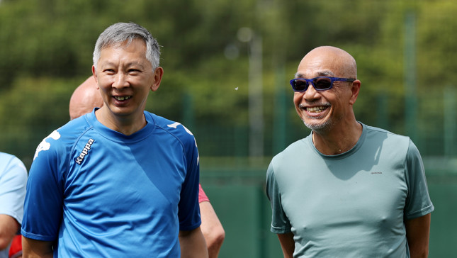 Two men stand outdoors on a sports field, one in a blue Kappa shirt and the other in a Quechua shirt. They are surrounded by trees and a blurred background.