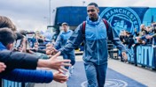 A Manchester City player in a tracksuit shakes hands with fans outside a stadium with the team's logo visible.