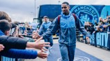 A Manchester City player in a tracksuit shakes hands with fans outside a stadium with the team's logo visible.