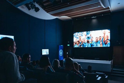 People seated in a theater watching a presentation about football on a large screen. The screen shows players celebrating a victory with a trophy.