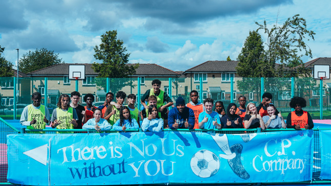 Group of children and adults standing on a soccer field with a banner reading 'There is No Us without YOU.' The banner has the C.P. Company logo and a soccer ball illustration.