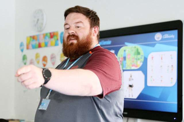 A bearded person with a red sleeve directs attention to a wall screen showing a Manchester City-themed presentation in a classroom.