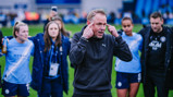 A blurred image of individuals celebrating on the field wearing Manchester City sports gear. The scene includes people taking pictures and a person gesturing energetically.