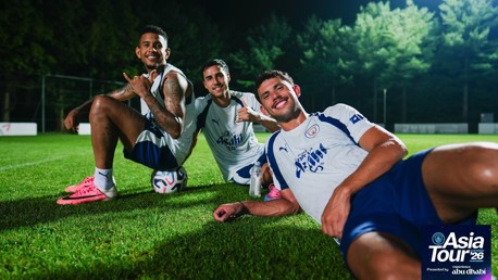 Three Manchester City players are on a football field at night, smiling and posing casually with a ball. The image features logos and text related to Asia Tour 2026 presented by Experience Abu Dhabi.