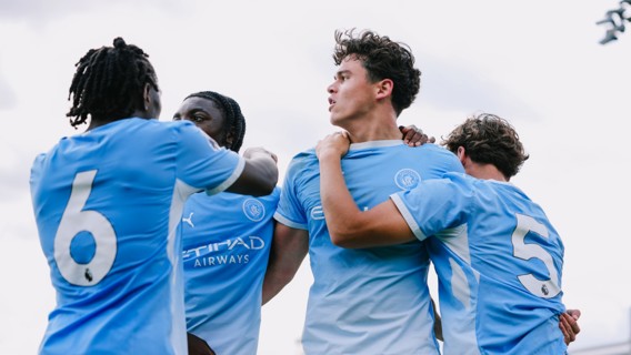 Young Manchester City players celebrating in blue jerseys, showing camaraderie after scoring. Visible jersey numbers are 6 and 5.