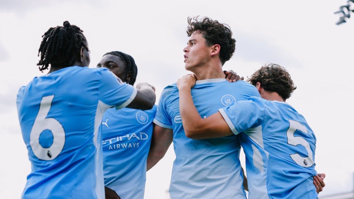 Young Manchester City players celebrating in blue jerseys, showing camaraderie after scoring. Visible jersey numbers are 6 and 5.