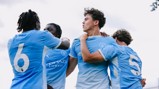 Young Manchester City players celebrating in blue jerseys, showing camaraderie after scoring. Visible jersey numbers are 6 and 5.