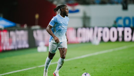 A soccer player from Manchester City in the club's light blue and white kit is actively running on a football field. A ball is nearby, and there is a blurred crowd in the background.