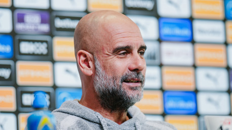 A person in a grey hoodie at a press conference with a Manchester City backdrop.