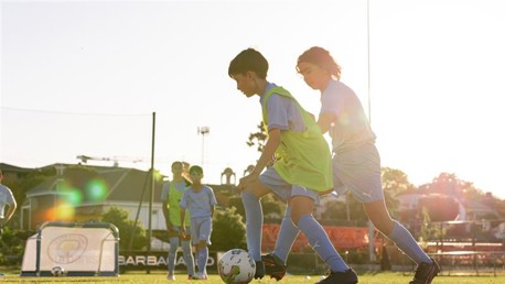 Children playing soccer on a field during sunset wearing blue uniforms and green bibs.