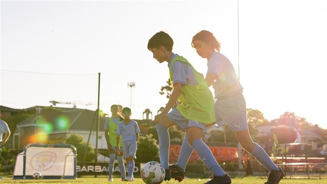 Children playing soccer on a field during sunset wearing blue uniforms and green bibs.