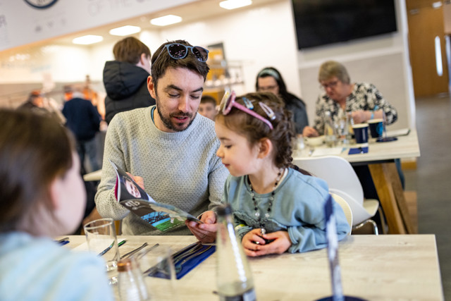 A man and a young girl sitting at a restaurant table, looking at a menu. Other people dining in the background.