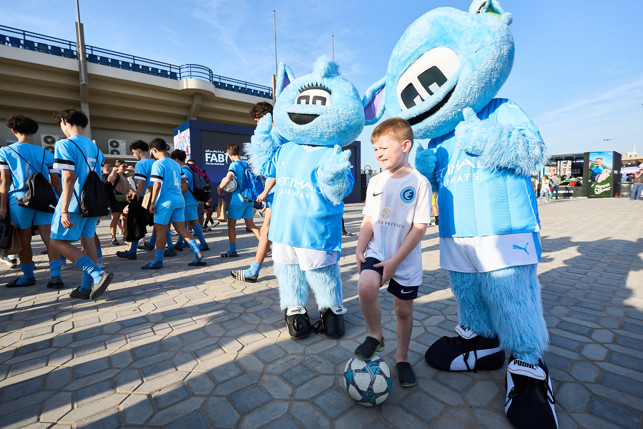 Two Manchester City mascots posing with a young fan in front of a football stadium. The young fan has one foot on a football. Other people in blue Manchester City outfits are in the background.