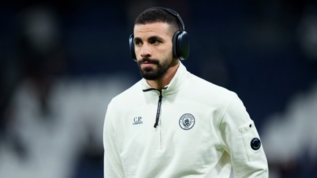 A Manchester City player in a white C.P. Company tracksuit with headphones before a match. The outfit has the Manchester City logo.