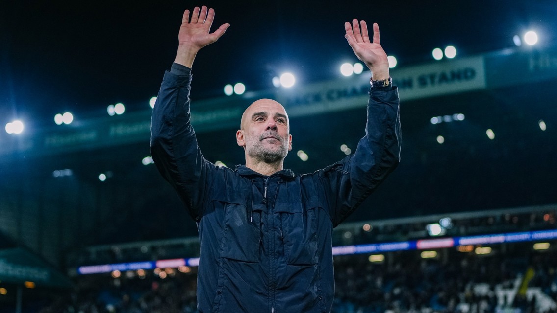 A person in a black jacket raises both arms while standing in a football stadium with bright lights in the background.