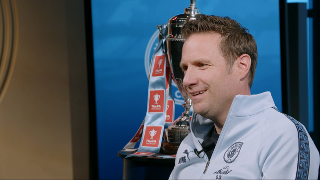 A person in a Manchester City tracksuit sits beside the FA Youth Cup trophy decorated with ribbons.