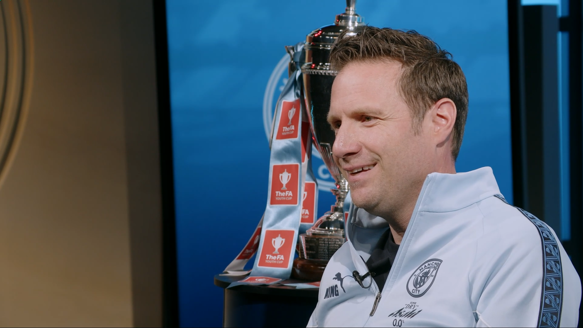 A person in a Manchester City tracksuit sits beside the FA Youth Cup trophy decorated with ribbons.
