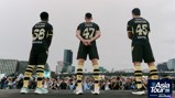 Three people in black Manchester City jerseys standing on a stage facing a large crowd with cameras, part of the Asia Tour '26 event.