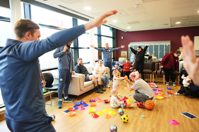 A group of adults and children, with some in Manchester City sports attire, interact and play games with various sports equipment in a community room.