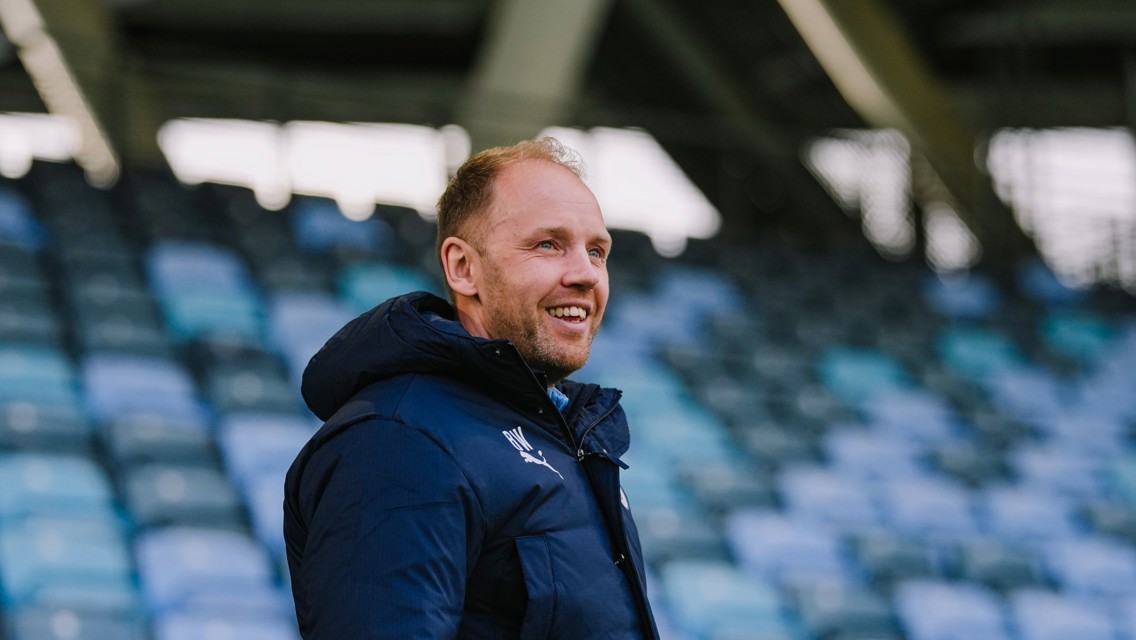 A person with a blurred face standing in an empty sports stadium wearing a blue Puma jacket.