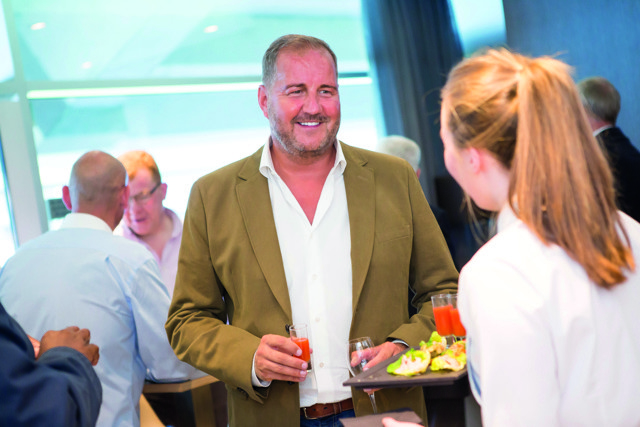 A man in a brown blazer holding drinks at a social gathering while conversing with a woman carrying a tray of food.