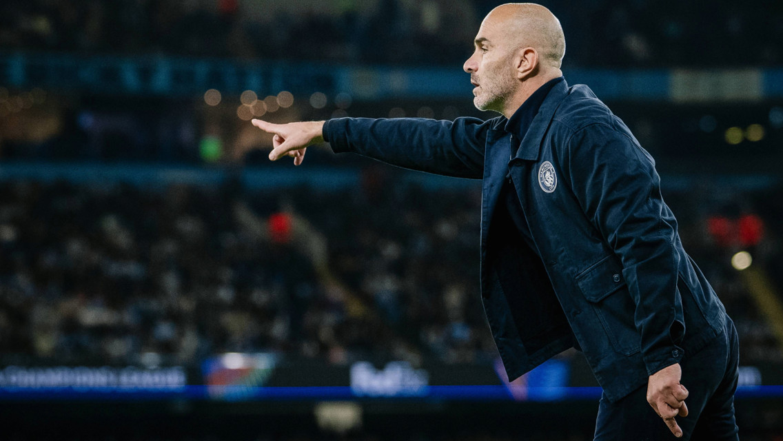 A football coach wearing a black jacket is pointing on a field during a match.