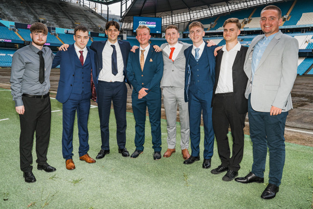 A group of eight young men in suits standing on the field inside a football stadium, identified as the Etihad Stadium, with faces blurred.