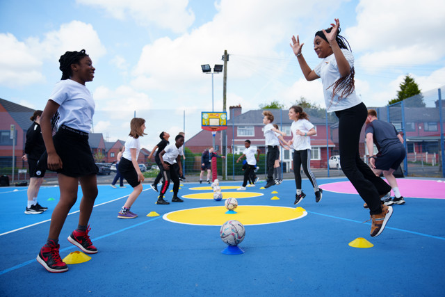 Group of children in white shirts and black shorts participating in an outdoor training session with soccer balls and cones on a blue court.