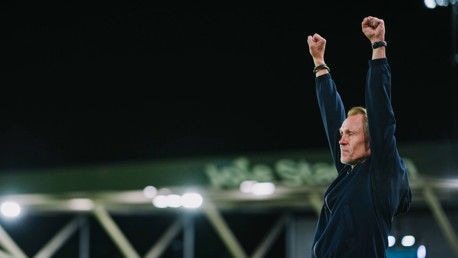 A person in a dark jacket is raising both arms in a victory gesture against a dark background.