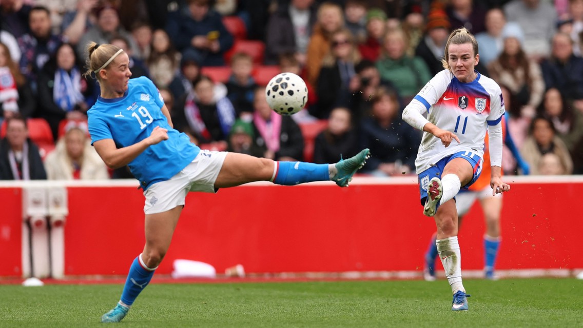 Two female soccer players in action during a match, with one player in a blue jersey and the other in a white jersey attempting to kick the ball.