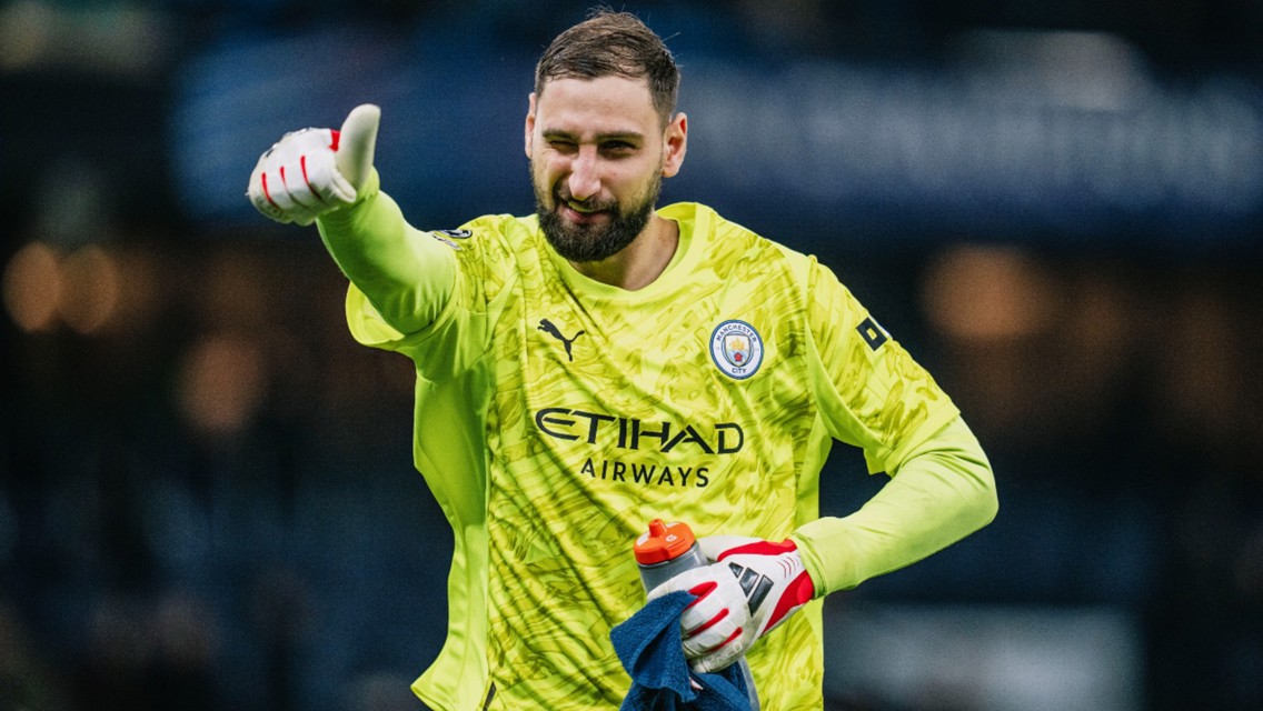 A goalkeeper wearing a neon green Manchester City kit, gloves and holding a water bottle on a soccer field.