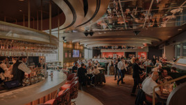 A modern restaurant interior with a curved bar counter, tables filled with diners, stylish chairs, and a mirrored ceiling.