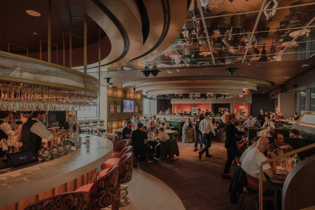 A modern restaurant interior with a curved bar counter, tables filled with diners, stylish chairs, and a mirrored ceiling.