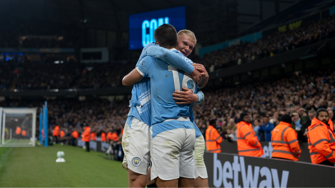 Manchester City players celebrating a goal at Etihad Stadium with a scoreboard showing 'GOAL' and security personnel in orange jackets visible.