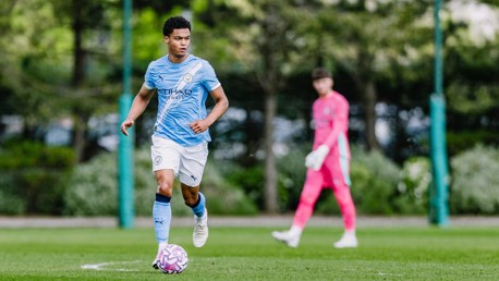 A football player in a Manchester City jersey dribbles the ball on a field, with a goalkeeper in the background.